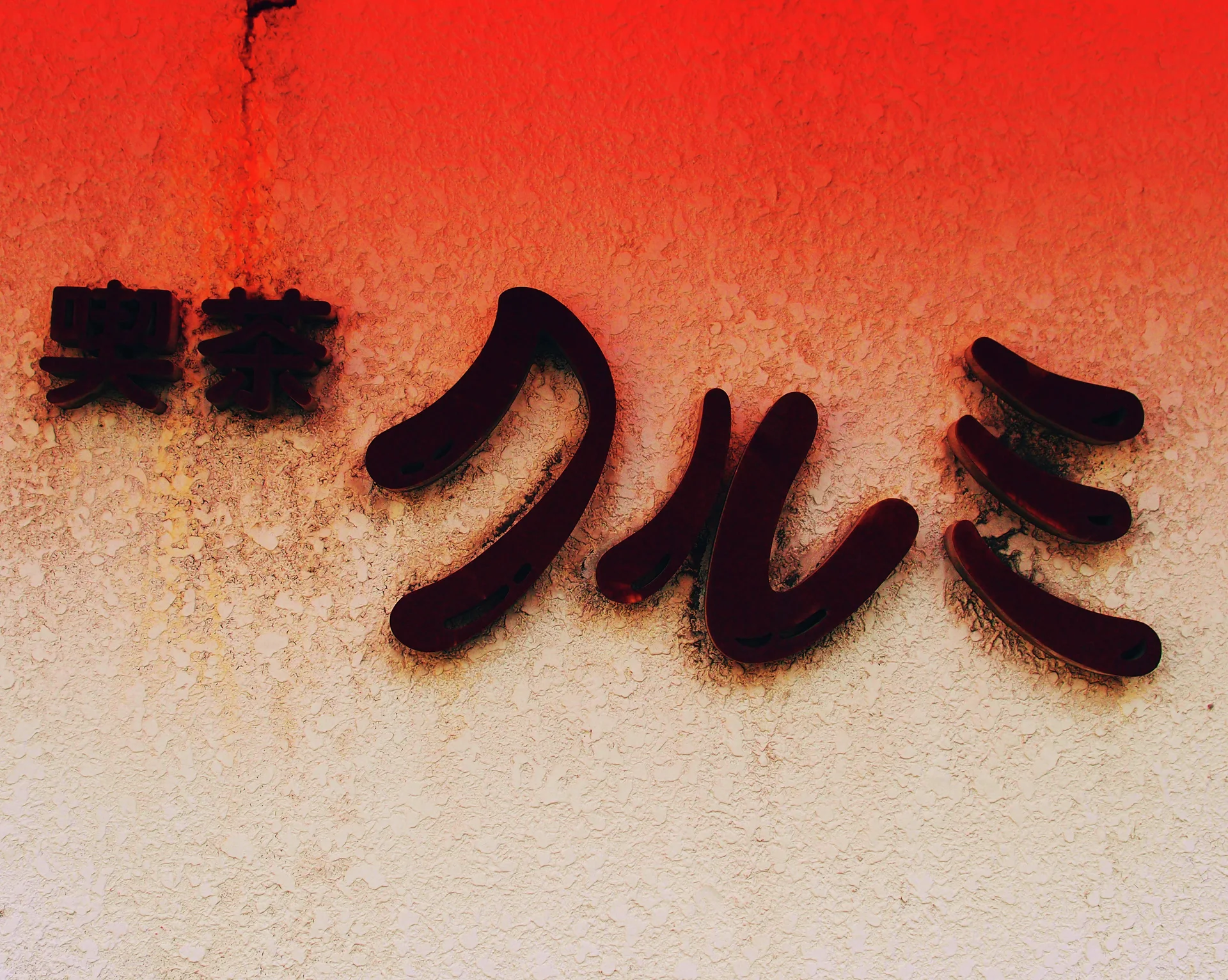 Raised dark Japanese characters cast shadows across a warm orange and cream textured wall.
