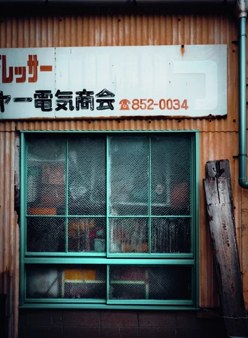 A faded business sign hangs above a green-framed window in a brown corrugated wall.
