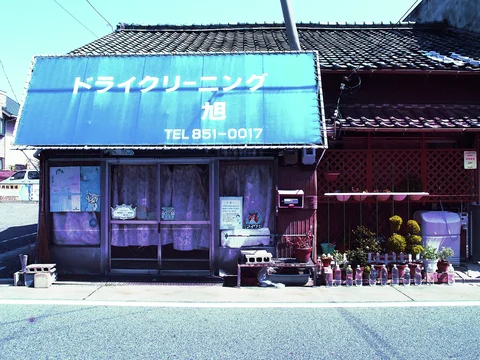 A blue Japanese awning shades weathered sliding doors beside rows of small plant pots beneath tiled roofs.
