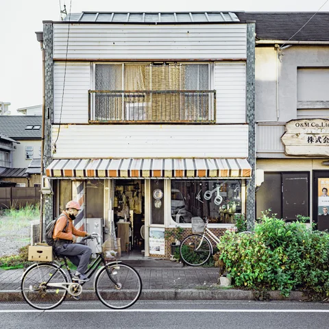 A cyclist rides past a narrow white storefront with a striped awning and leafy plants.