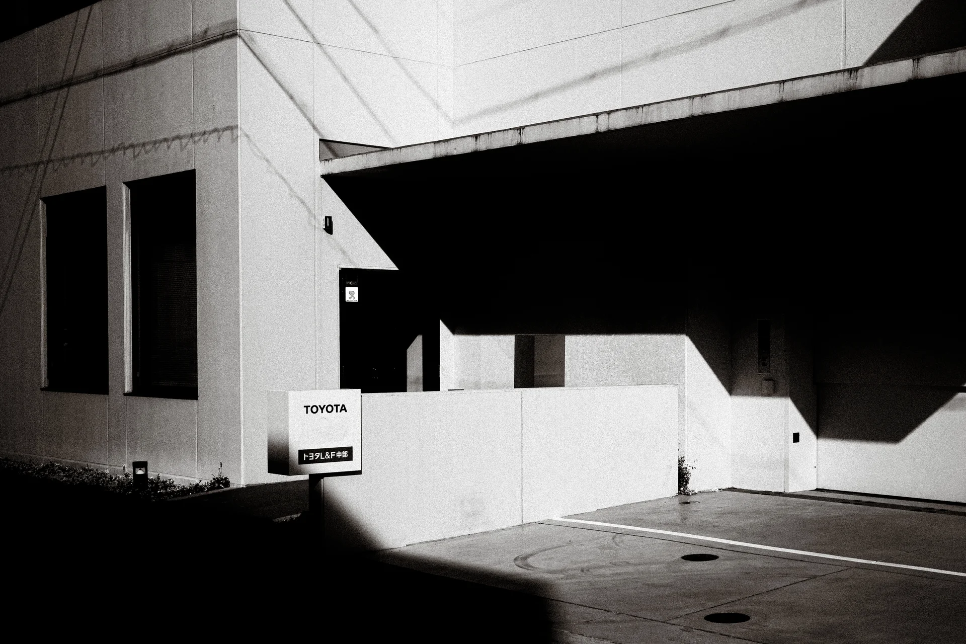 Sharp angular shadows cross a pale building and empty marked parking spaces in black and white.