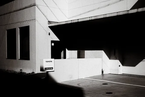 Sharp angular shadows cross a pale building and empty marked parking spaces in black and white.
