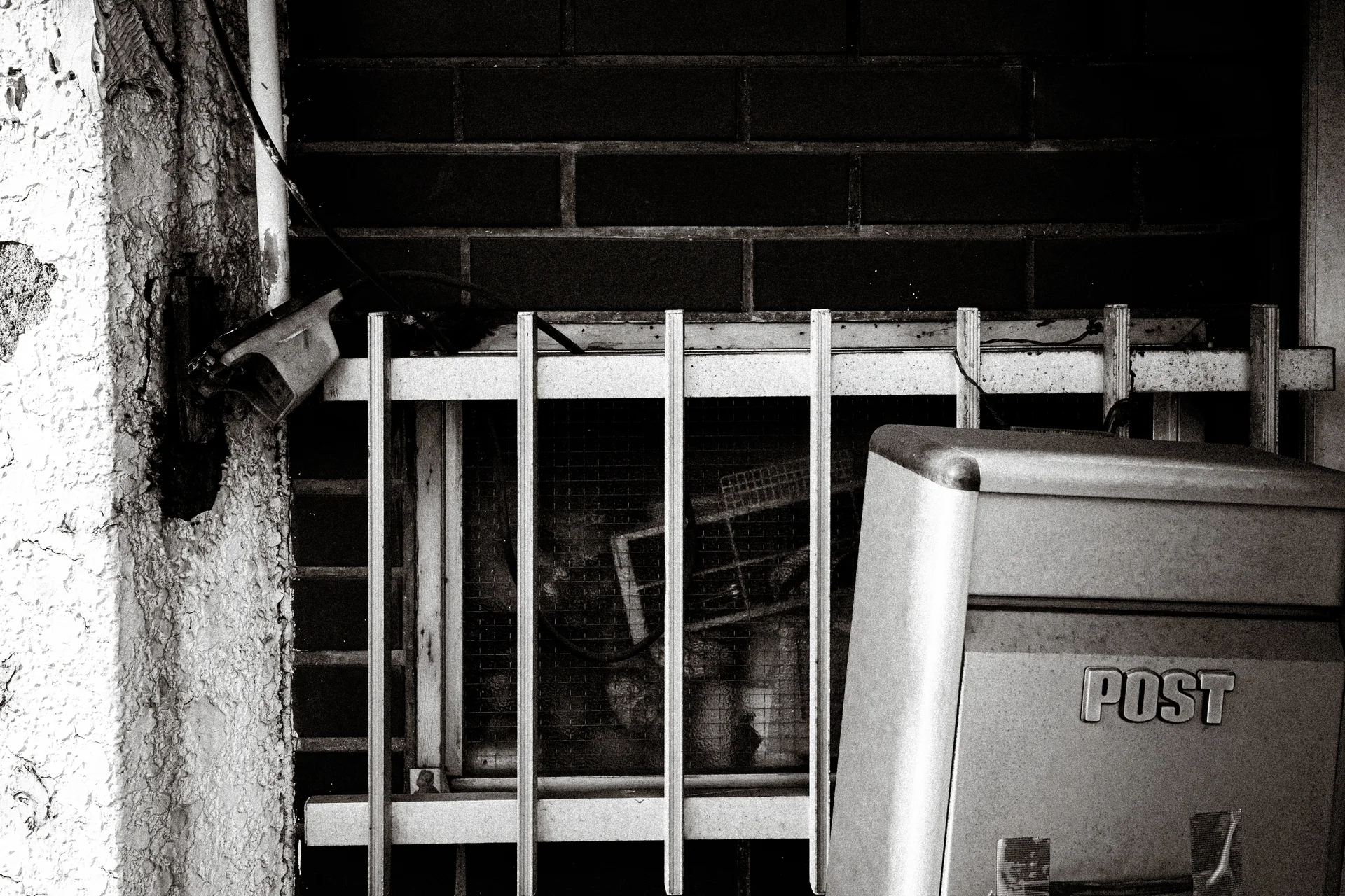 A metal mailbox stands before a barred window and dark brick wall in a black and white close-up.