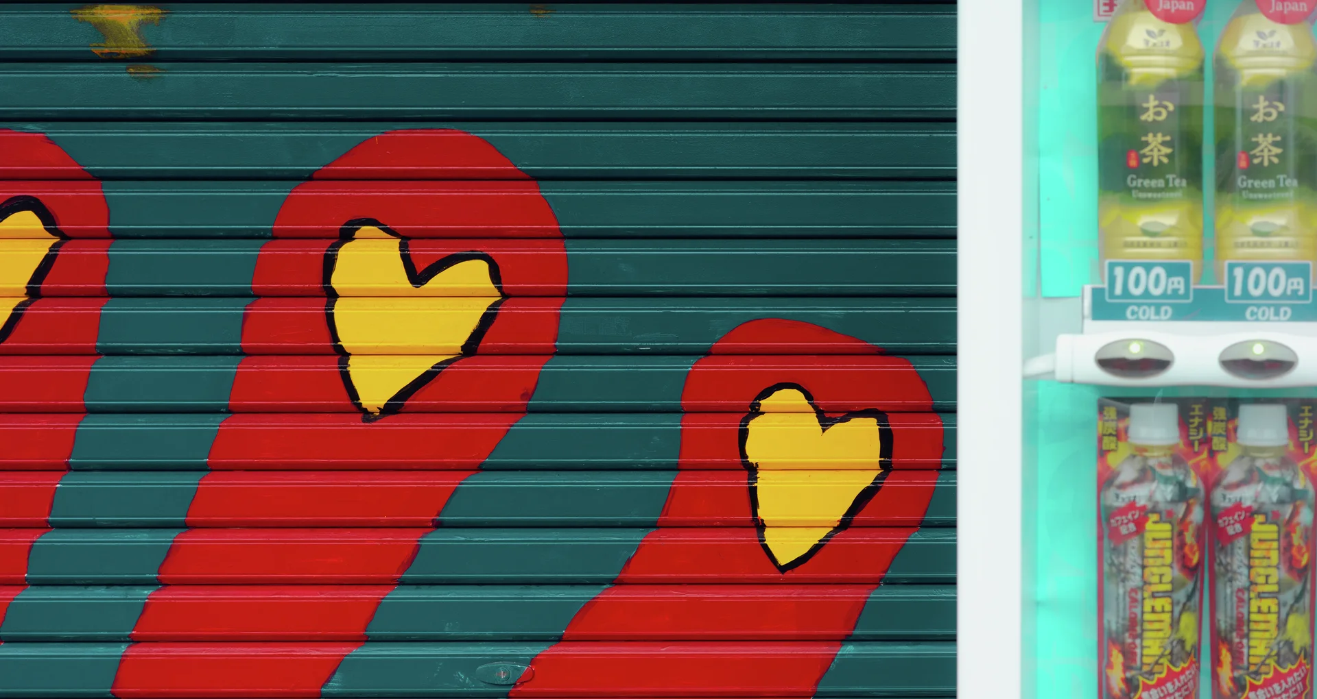 Yellow hearts decorate red shapes on a dark green shutter beside a drink vending machine.