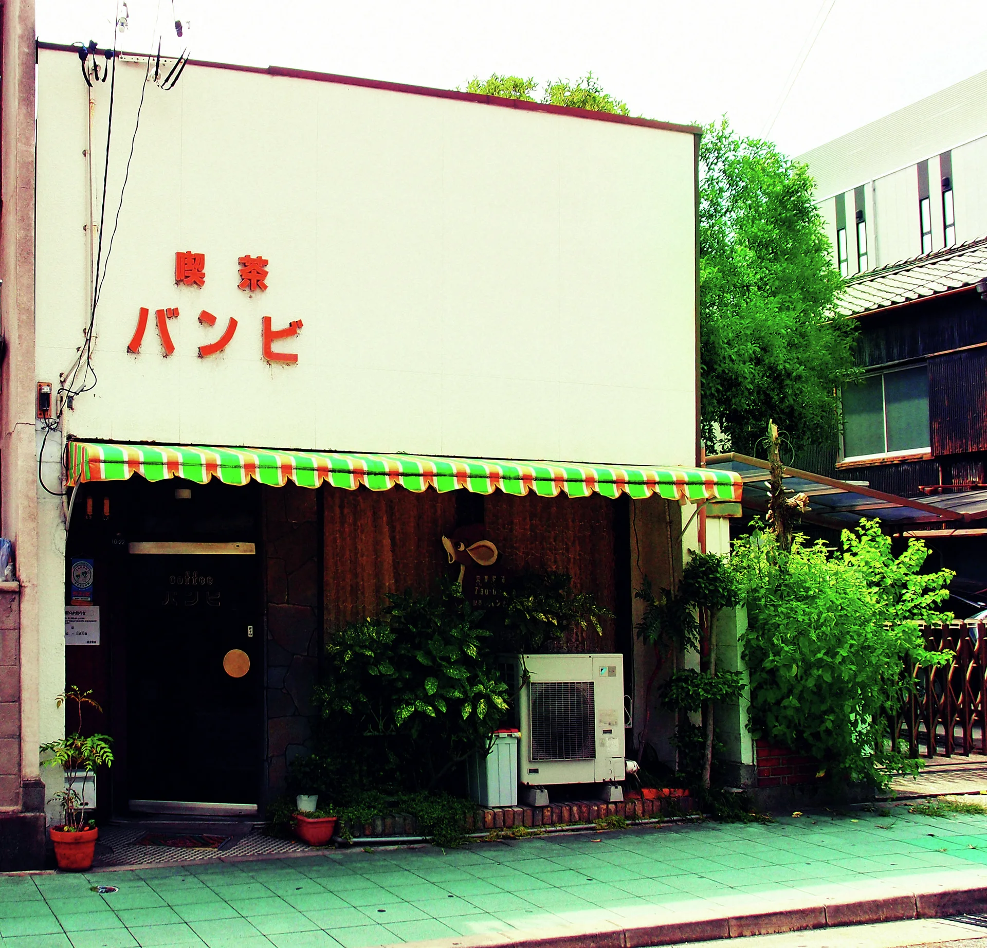 Red Japanese lettering decorates a pale cafe facade above a green-striped awning and shaded entrance.