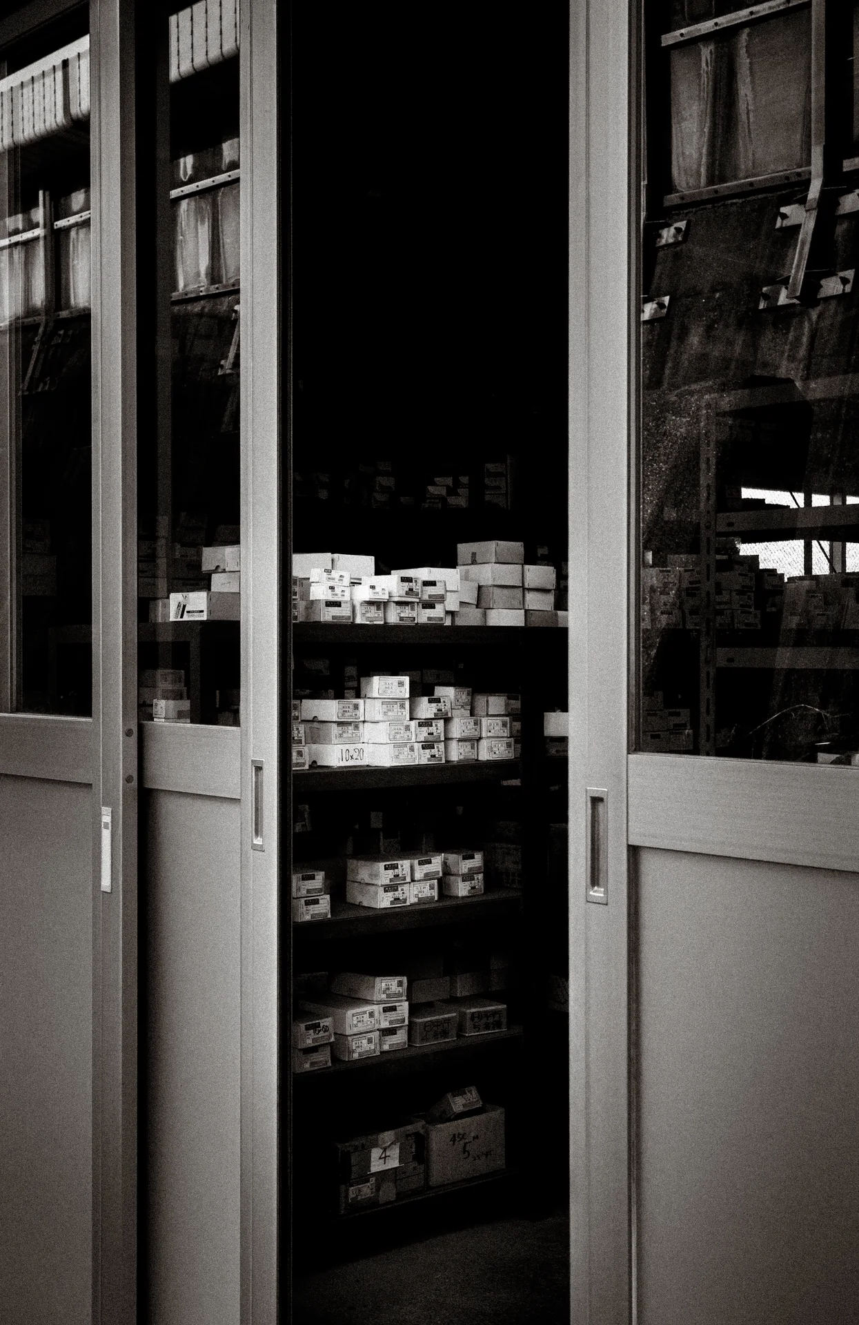 Partly open glass doors reveal shelves of small boxes inside a dark shop in black and white.