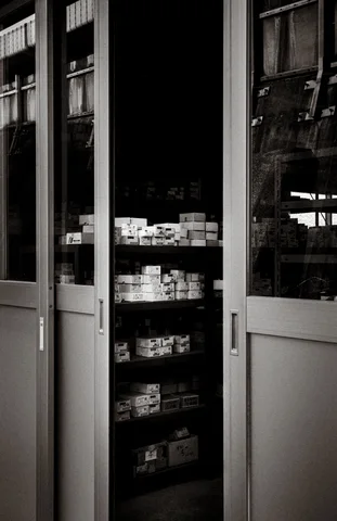 Partly open glass doors reveal shelves of small boxes inside a dark shop in black and white.
