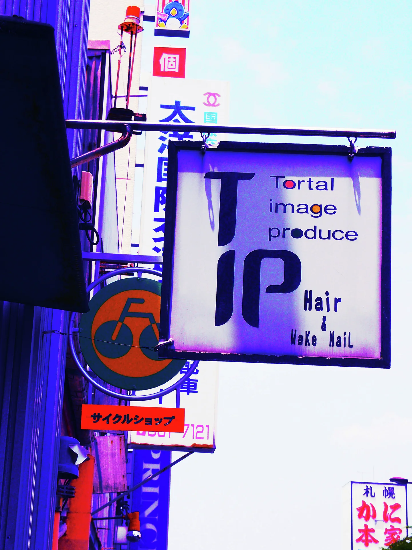 A square hair salon sign hangs among vertical shop signs against a pale blue sky.
