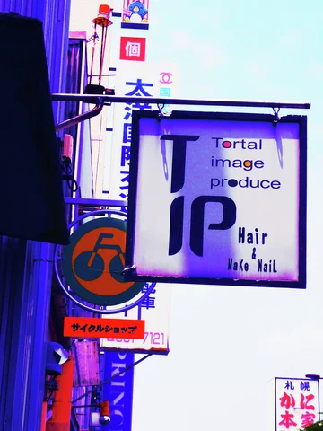 A square hair salon sign hangs among vertical shop signs against a pale blue sky.