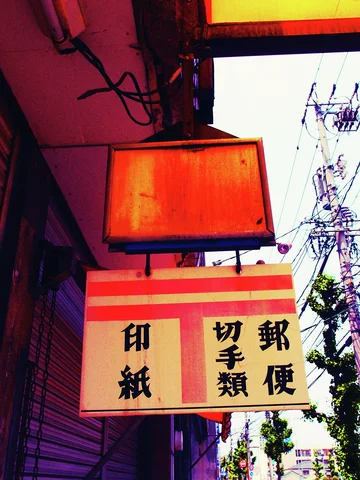 Two rectangular hanging signs project over a shuttered storefront beside utility wires and a leafy tree.