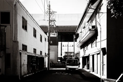 A small truck sits in a narrow lane between buildings beneath an elevated structure in black and white.
