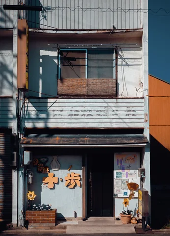 A narrow weathered storefront has orange characters, a boarded upper window, and small potted plants.