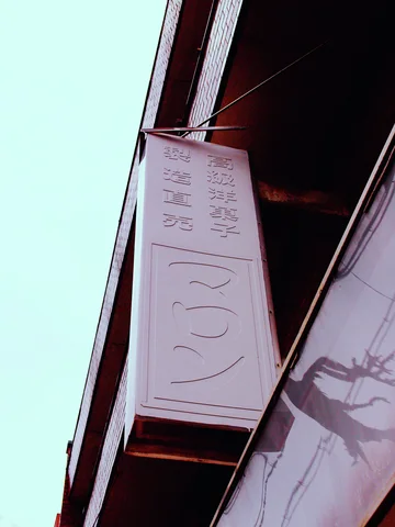 A tall pale projecting sign carries faint embossed characters beside a dark building edge and blue sky.