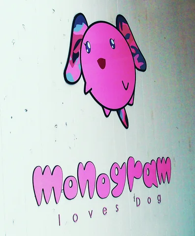 A pink cartoon dog decorates a pale sign above rounded Monogram lettering and smaller black text.