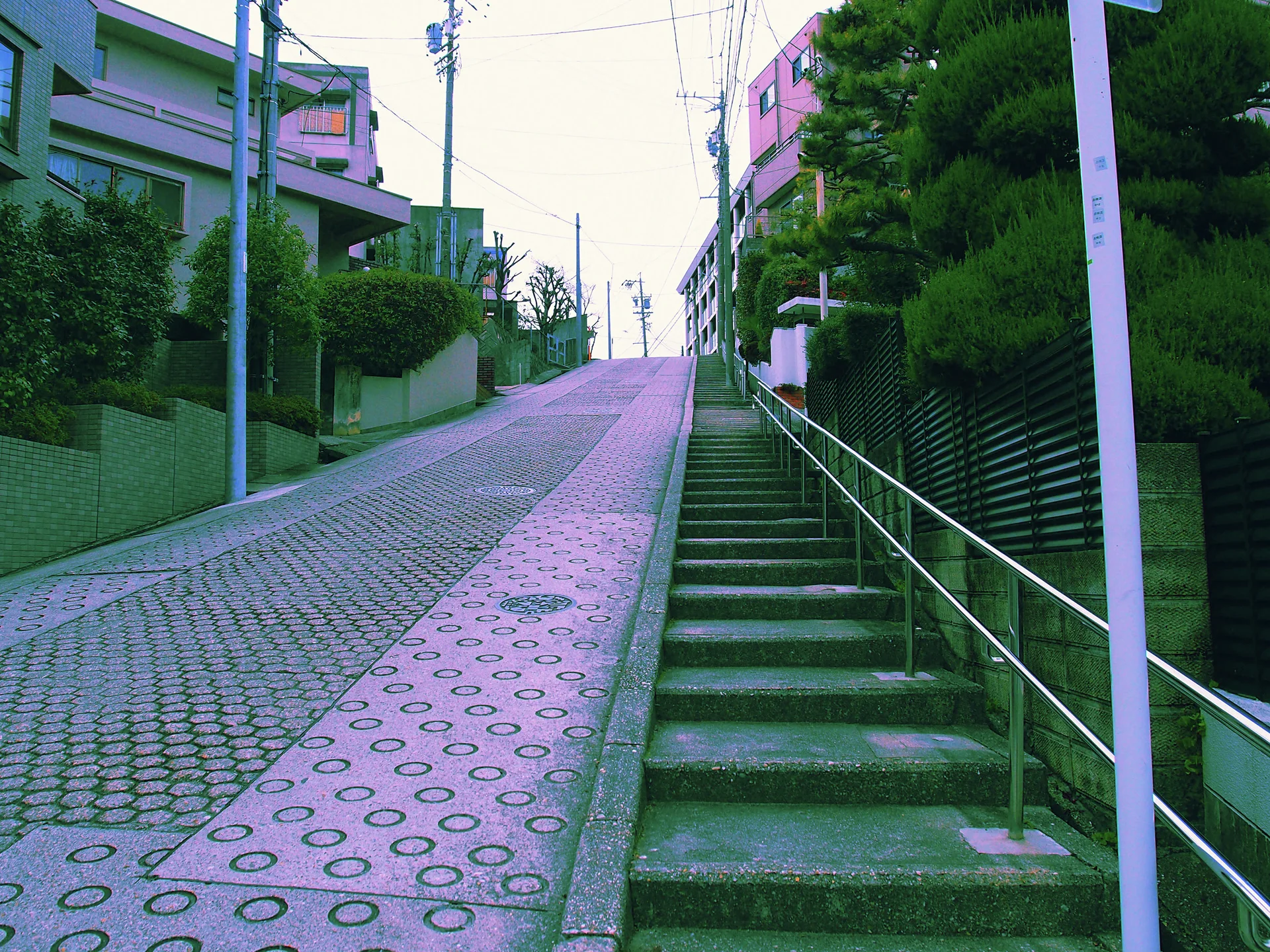A steep textured lane climbs beside a long concrete staircase with metal handrails.