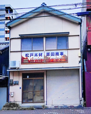 A narrow pale building has blue upper shutters and a sign above a closed ground-floor shutter.