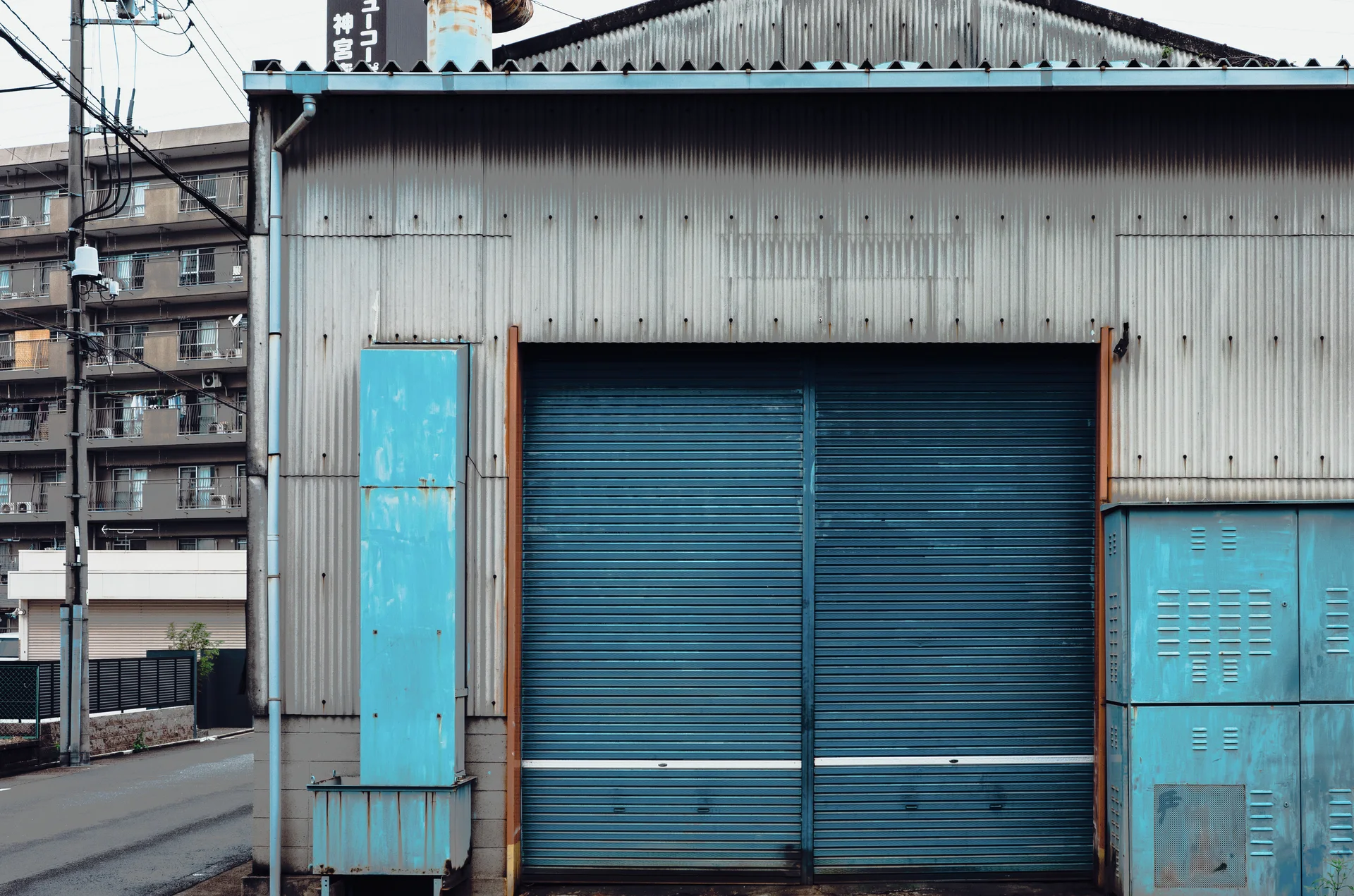 Blue shutters and matching worn panels mark a corrugated warehouse facade beside a narrow road.