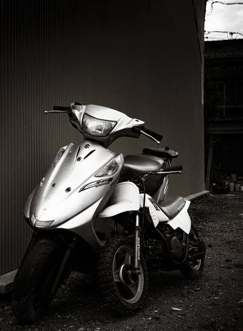 A pale scooter stands against a dark corrugated wall on rough ground in black and white.