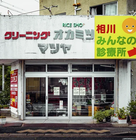 Raised Japanese lettering and a bright yellow sign sit above glass shop doors and small potted plants.