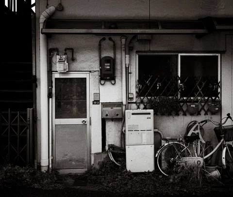Bicycles stand beside a small doorway beneath exposed pipes and a barred window in black and white.