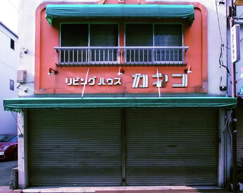 A salmon-colored storefront has closed green shutters, turquoise awnings, and a small balcony above raised Japanese lettering.