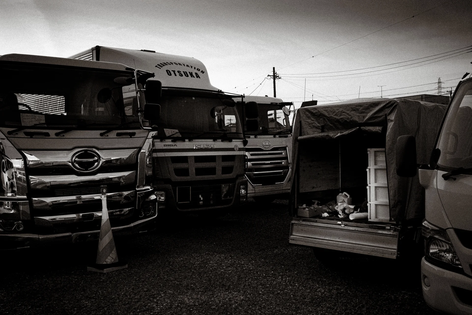 Several large trucks crowd a gravel yard beside an open tarpaulin-covered vehicle in black and white.