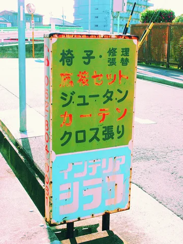 Multicolored lettering fills a narrow weathered freestanding sign beside a pale sidewalk.
