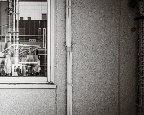 A geometric window grille reflects nearby buildings beside a textured wall and drainpipe in black and white.