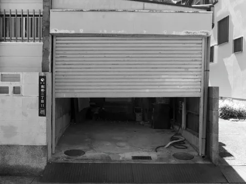 A partially raised metal shutter reveals a dark garage with exposed pipes and rough flooring in black and white.