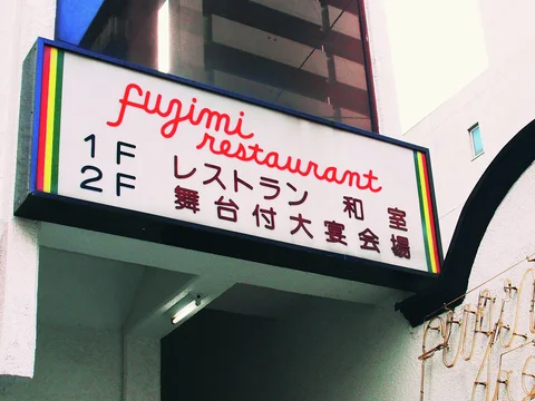 Red cursive Fujimi Restaurant lettering fills a white projecting sign edged with narrow rainbow stripes.