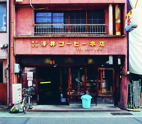 Gold Japanese lettering decorates a reddish coffee shop facade above a dark entrance and parked bicycle.