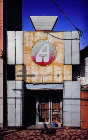 A rust-streaked metal facade has a faded circular emblem above a narrow entrance and tiled pavement.