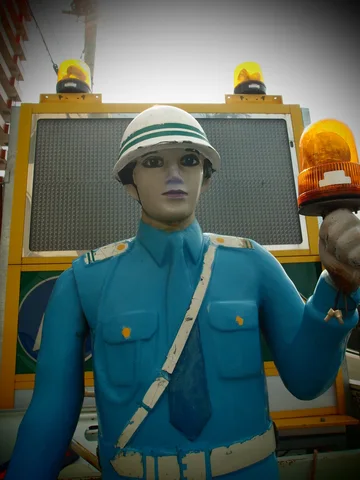 A mannequin in a blue uniform and white helmet holds an amber beacon before a yellow-framed panel.