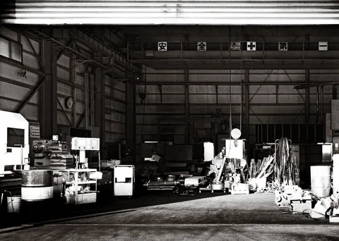 Machinery, barrels and piled metal pieces fill a broad open warehouse bay in black and white.