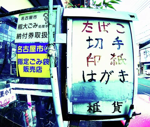 A faded vertical shop sign with multicolored characters stands beside smaller blue and yellow notices.