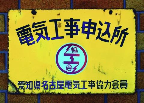 Dark blue Japanese lettering and a circular emblem fill a weathered yellow plaque mounted on brick.