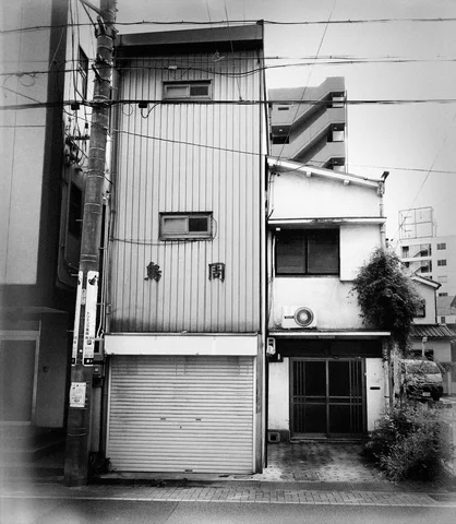 A narrow corrugated building with tiny windows and a closed shutter stands beside an older house.