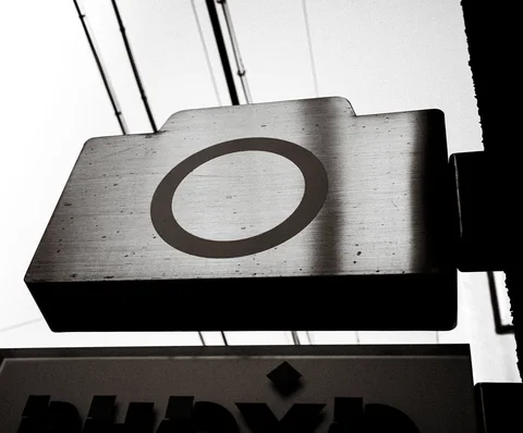 A projecting rectangular sign bears a large dark ring against a pale sky in black and white.