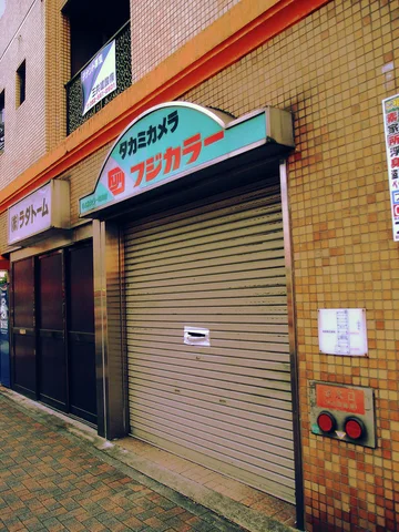 A curved pale blue sign tops a closed shutter on a yellow-tiled storefront beside a brick sidewalk.