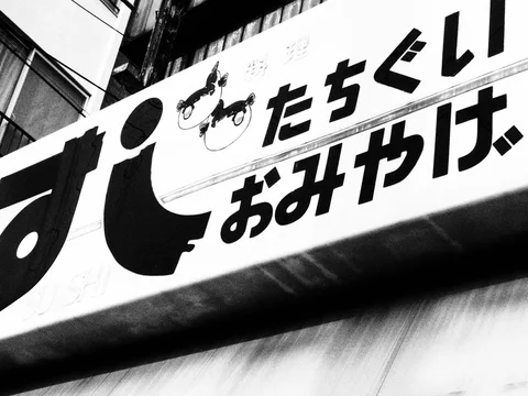 Bold Japanese characters and two illustrated faces span a diagonal shop sign in black and white.