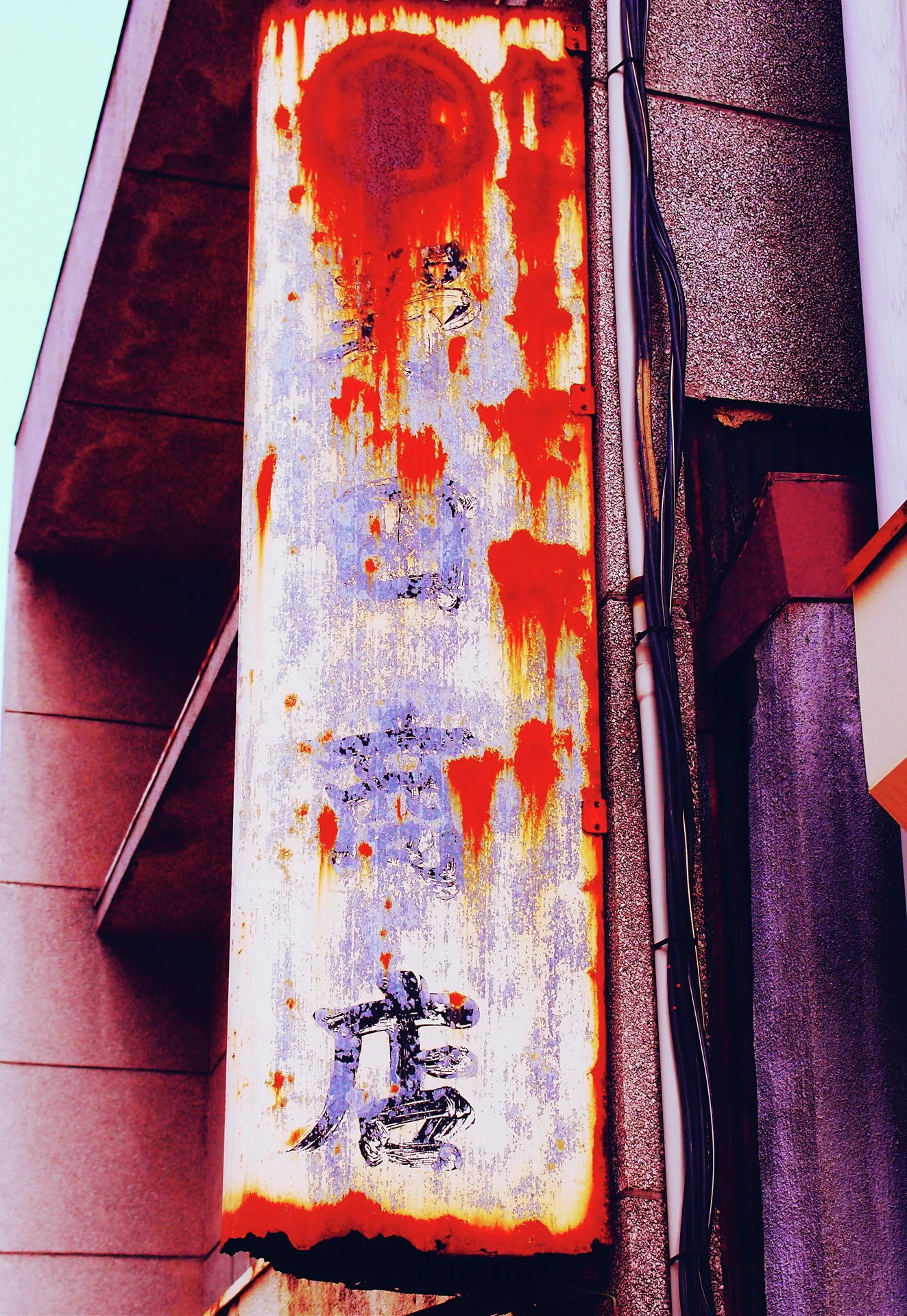 Heavy red rust streaks obscure faded lettering on a tall white sign beside a building wall.