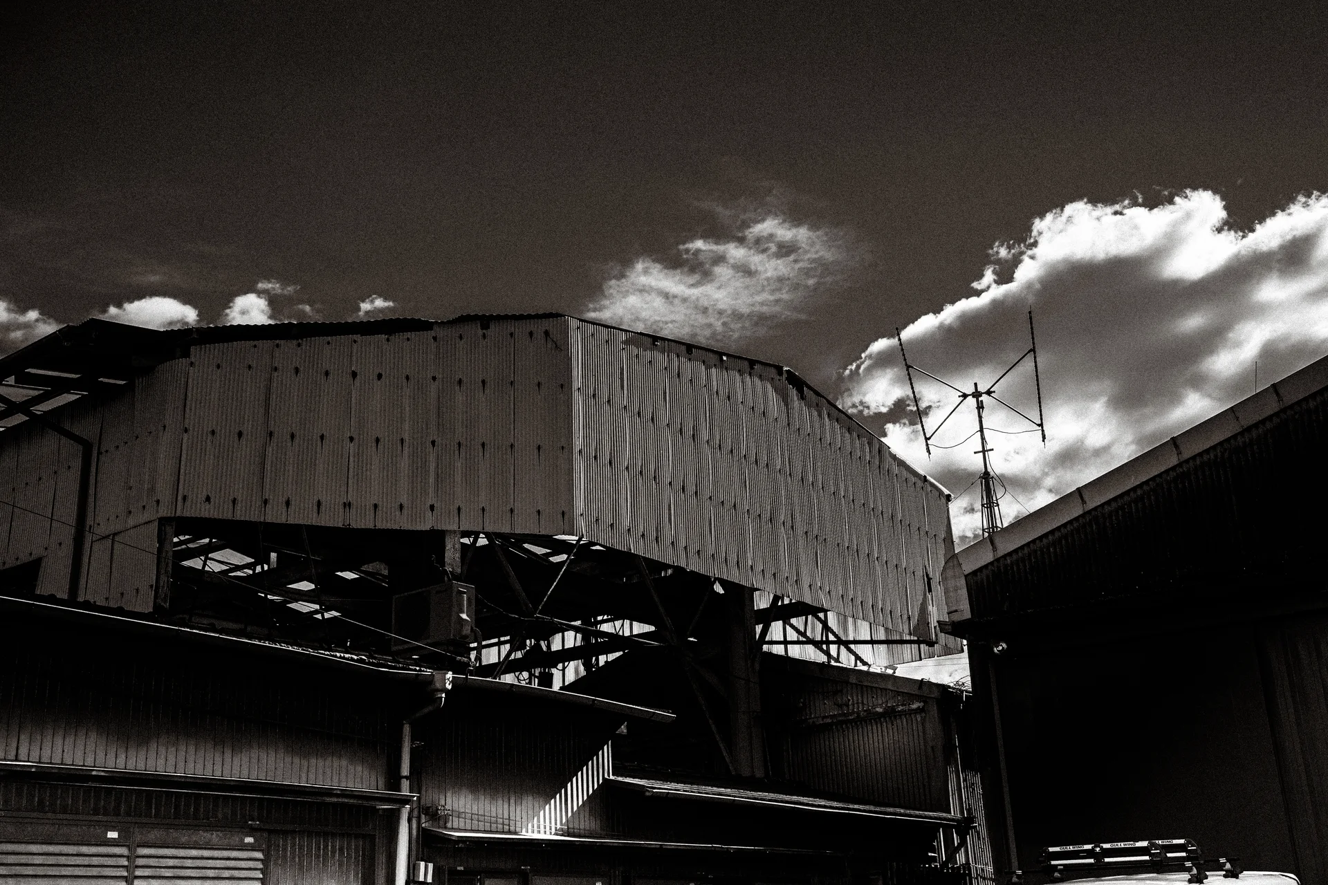 Corrugated industrial roofs and exposed steel beams rise beneath bright clouds in black and white.