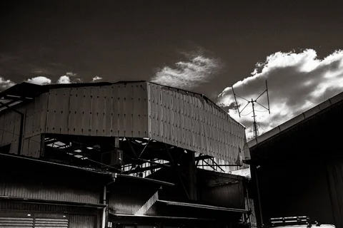 Corrugated industrial roofs and exposed steel beams rise beneath bright clouds in black and white.