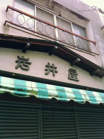 A green and white striped awning shades a closed shutter beneath Japanese lettering and a circular-patterned balcony railing.