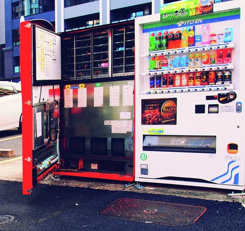 A red cabinet stands with its door open beside a white drink vending machine on dark pavement.