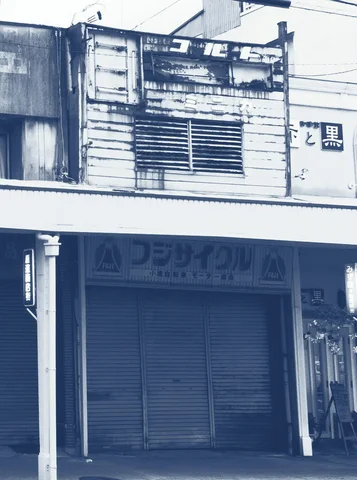 A weathered white shop facade has dark upper panels above a closed shutter and faded signage.