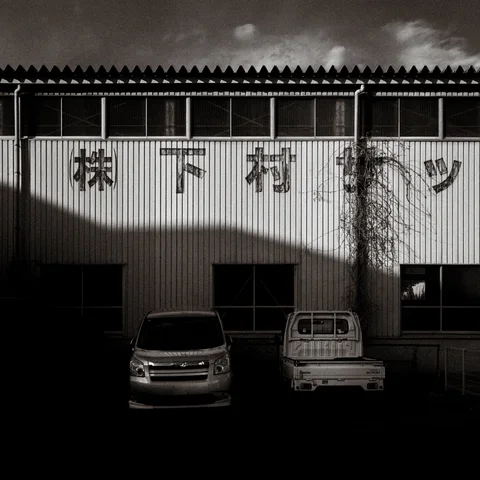 Two vehicles stand outside a corrugated warehouse crossed by broad shadows in black and white.