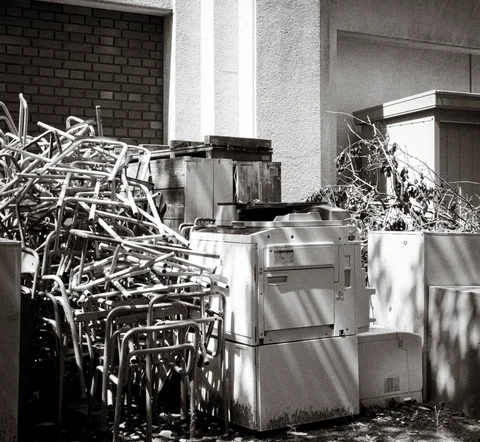 Stacks of metal frames and discarded appliances crowd a sunlit building corner in a black and white view.