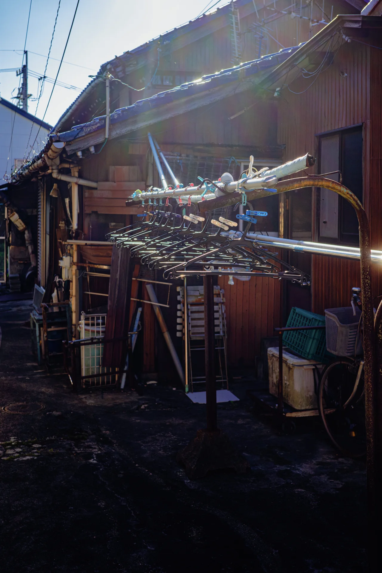 Metal laundry racks crowd a shadowed yard beside a weathered wooden house with sunlight along its roof.