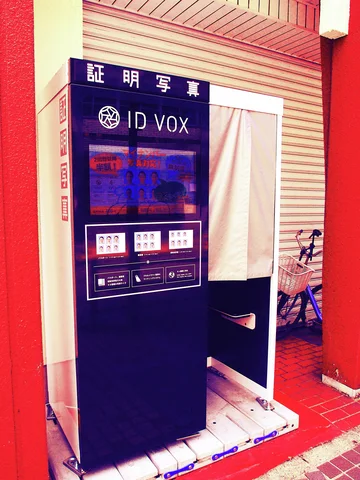 A dark identification photo booth stands against a closed shutter beneath red and white trim.